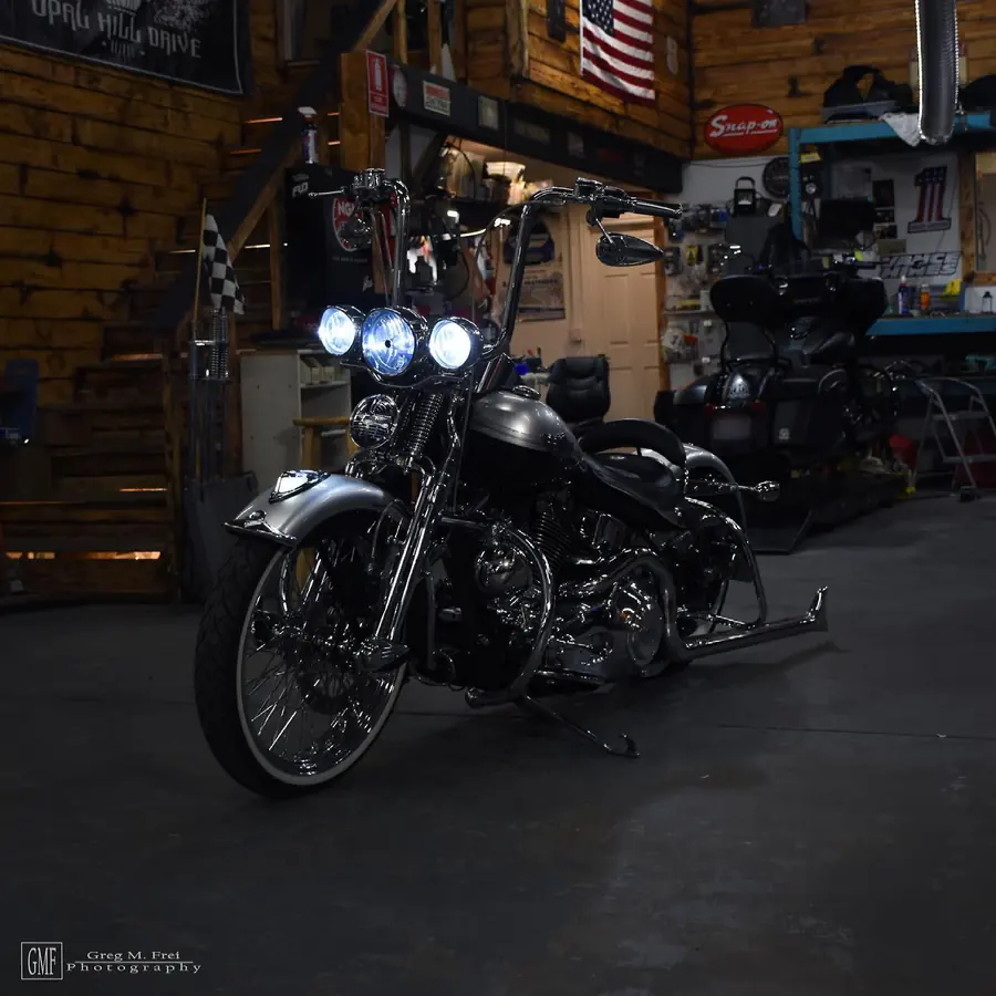 Silver Softail with triple headlights, tall bars, and chrome fishtail pipes in a wood-paneled shop